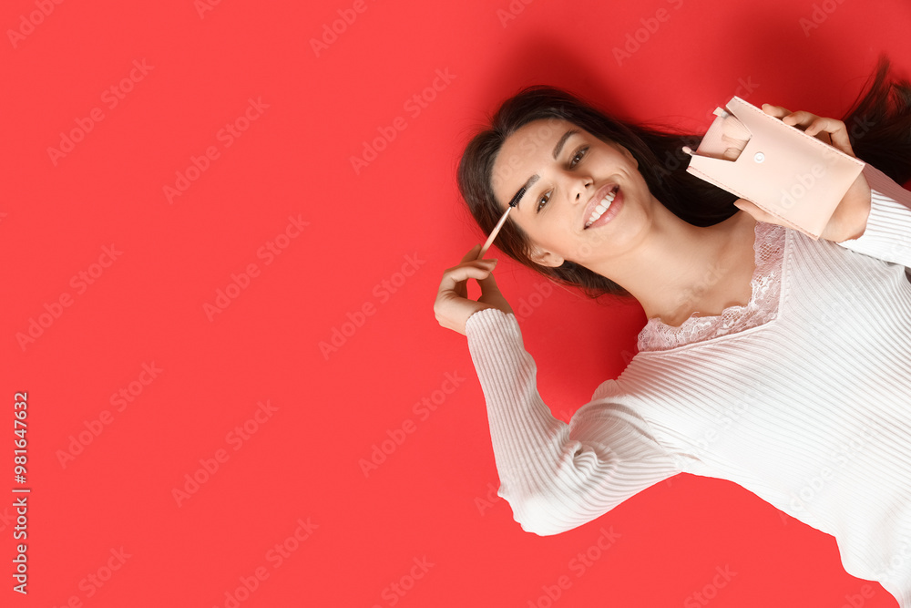 Happy smiling young woman with makeup brushes lying on red background