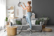 © Pixel-Shot - Young woman on stepladder screwing light bulb into hanging lamp at home