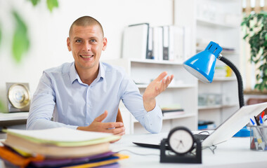 Portrait of polite smiling entrepreneur sitting at table in office, making welcome gesture. Invitation to negotiate concept