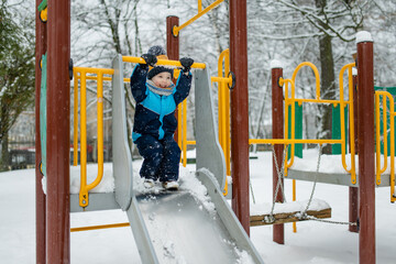 Wall Mural - Adorable little boy having fun on a playground on snowy winter day. Cute child wearing warm clothes playing in a snow.