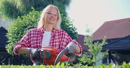 Wall Mural - Smiling young woman with blond hair cutting bushes with electric trimmer. Female gardener in safety glasses and gloves shaping overgrown hedge outdoors.