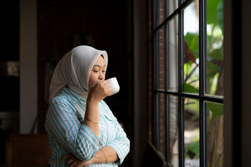 A woman wearing hijab and holding a cup of coffee. A woman wearing a scarf and holding a cup of coffee. She is smiling and looking out the window
