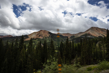 Photo of mountains and pine trees blue sky with white fluffy clouds taken from moving car