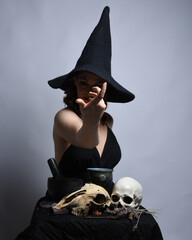 Poster - Close up portrait of beautiful  female model wearing black dress, pointy witch hat halloween costume.  Isolated figure on studio background. Casting magic spell at spooky occult  ritual potion table 