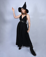 Wall Mural - portrait of beautiful female model wearing glamorous black witch dress, pointy hat halloween costume. walking pose holding broomstick, isolated figure on studio background