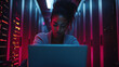 © Hikmet - An engaging illustration of a Black woman IT support engineer working diligently in a dark server room filled with servers and network equipment. She is focused on her laptop, showcasing her expertise