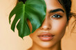 © Hanna - Young woman with a striking gaze partially covered by a tropical leaf against a neutral background in a bright indoor setting