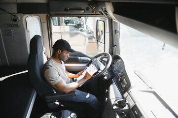 African-American truck driver is sitting behind the wheel holding a clipboard and checking the documents for the cargo