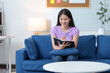 © crizzystudio - Young Asian woman sitting on the sofa in the living room at home. Relaxing, reading a book, keeping a diary happily. Fresh in the morning Lifestyle concept, hobbies