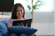 © crizzystudio - Young Asian woman sitting on the sofa in the living room at home. Relaxing, reading a book, keeping a diary happily. Fresh in the morning Lifestyle concept, hobbies
