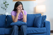 © crizzystudio - A woman with a sneeze is feeling unwell. Runny nose and sneezing, stomach ache, sore throat, fatigue in the living room on the sofa in the house.  Feeling bad and having allergies treatment concept.