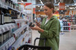 © Grustock - Young woman shopping in a store, holding a jar of cosmetics while comparing prices through a smartphone app, making informed decisions on beauty products and promotions