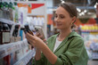 © Grustock - Young woman shopping in a store, holding a jar of cosmetics while comparing prices through a smartphone app, making informed decisions on beauty products and promotions