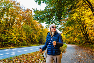 Wall Mural - Portrait of middle-aged blonde woman in casual wear walking with poles on sidewalk by street in autumn scenery. Autumnal Nordic walking in city.