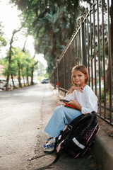 Smartphone in hand, sitting. Schoolgirl with backpack is outdoors