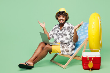 Wall Mural - Full body young IT Indian man wear beach shirt hat rest near hotel pool sit in deckchair work hold use laptop pc computer isolated on plain green color background. Summer vacation sea sun tan concept.