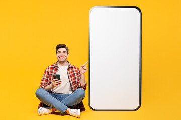 Wall Mural - Full body young man he wearing red checkered shirt casual clothes sit point on big huge blank screen mobile cell phone with area use smartphone isolated on plain yellow background. Lifestyle concept.