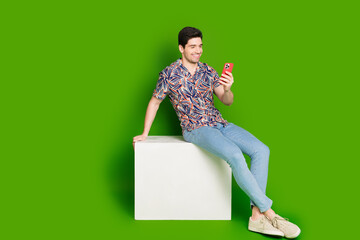 Sticker - Full size photo of positive guy dressed print shirt jeans sit on white platform look at smartphone isolated on green color background