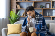 © Tetiana - Young man sitting on sofa covered with blanket, drinking hot beverage and blowing nose, feeling unwell and cold