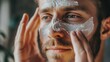 © fotofabrika - A young man applies a soothing facial mask in a well-lit room, focusing on skin care for relaxation and rejuvenation