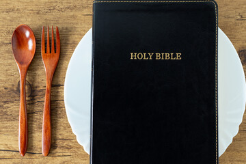 Holy bible book on empty plate with fork and spoon on wooden table. Top view. Christian fasting and spiritual food, biblical concept.