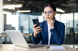 © Liubomir - Asian businesswoman in office using smartphone while working on laptop. She wears glasses and blue suit, appearing focused and professional. Image conveys productivity, technology, modern work