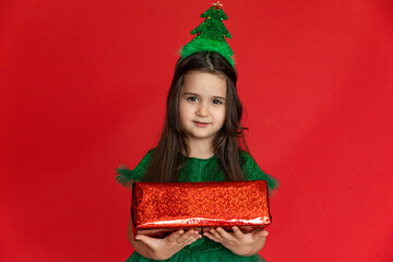 Happy little girl in a festive carnival costume of little Christmas tree with Christmas gift on red background. A little girl holds a gift on a red isolated background.