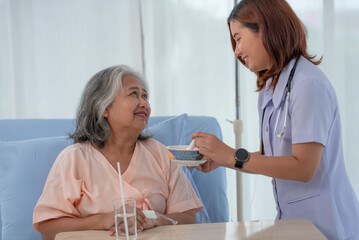 Asian nurse feeding breakfast to elderly female patient on bed in hospital ward