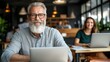 © angel_nt - A senior man with glasses and a beard sits at a laptop, smiling confidently in a contemporary workspace, as a young woman engages with her computer nearby