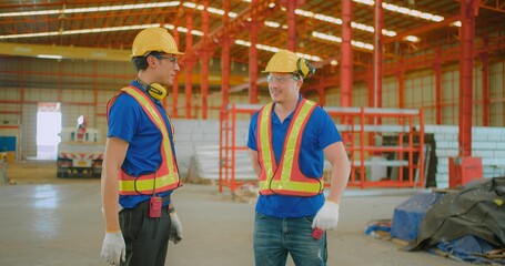 Two construction workers in safety gear talking inside a modern industrial warehouse with red steel framework and various construction materials.