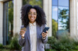 © Liubomir - Hispanic businesswoman standing outside modern building holds smartphone and credit card with bright smile. Represents professional success, digital presence, and business connectivity