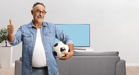 Sticker - Smiling mature man holding a football and gesturing thumbs up in front of tv