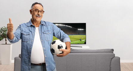 Poster - Smiling mature man holding a football and gesturing thumbs up in front of tv and sofa