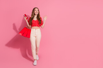 Poster - Full body photo of teen woman look empty space walk wave hand shopping device dressed stylish red clothes isolated on pink color background