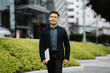 © Kritdanai - Confident Businessman Walking in the City: A young Asian man, dressed in a smart blazer and carrying a laptop, walks confidently through the urban landscape, radiating ambition and success.  His smile