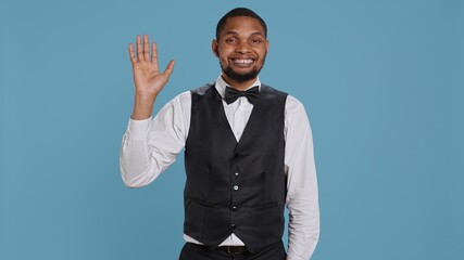 Bellboy employee smiling and waving hello at hotel guests, greeting people with confidence and offering luxury services for a pleasant stay. Cheerful personnel saying hi to people. Camera B.