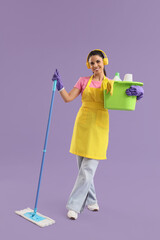 Canvas Print - Female janitor in headphones with cleaning supplies on lilac background