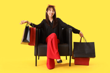 Wall Mural - Young Asian woman with shopping bags sitting in armchair on yellow background. Black Friday Sale