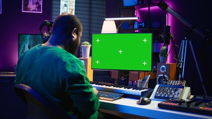 Producer composing music with electronic keyboard notes and green screen display on pc, post production in home studio. Musician artist plays midi controller next to mockup layout. Camera B.