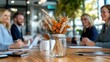 © Lens Legacy - A close-up of a glass jar vase filled with dried flowers and wheat stands on a wooden meeting table, with blurred professionals discussing in the background.