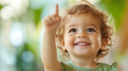 A joyful toddler with curly hair points upward, smiling brightly against a lush, green blurred background, capturing a moment of innocence and wonder.