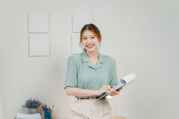 Confident Young Professional: A young Asian woman in stylish business casual attire stands confidently in her home office, holding documents and smiling brightly at the camera