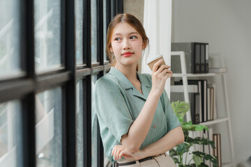 Coffee Break Contemplation: A young woman in a mint green blouse leans against a window, gazing thoughtfully out, a cup of coffee in hand. The natural light casts a warm glow, creating a serene and co