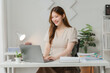 © Kritdanai - Focused Productivity: A young Asian woman beams at her laptop, engrossed in work at her bright, minimalist home office.