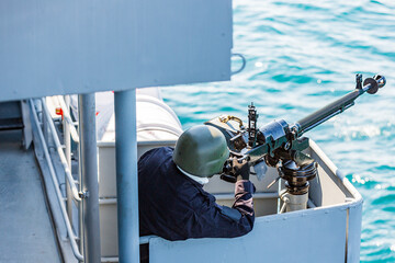 machine gun on a battleship. A marine holds a weapon. securing a ship. protection from pirates. a sailor in a black uniform is on automatic weapons duty on a battleship.