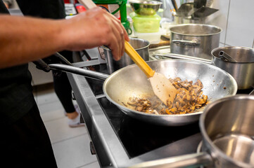 Close-up of a person cooking in kitchen, stirring mushrooms in pan with a wooden spatula. The scene captures the essence of home cooking, emphasizing the fresh ingredients and the culinary process. 