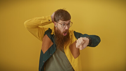 Shocked bearded redhead man in casual attire looking at his watch with a yellow background.