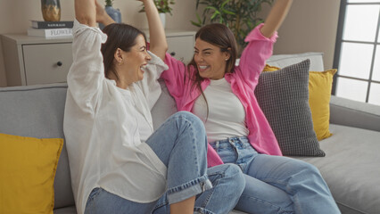 Two smiling women wearing casual outfits, socializing and celebrating together on a comfortable sofa in a cozy living room.