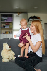 Happy Caucasian mum and baby sitting on the sofa at home looking at something with interest.