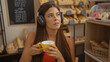 © Krakenimages.com - Beautiful hispanic young woman with headphones drinks coffee in a bakery shop interior with various pastries displayed